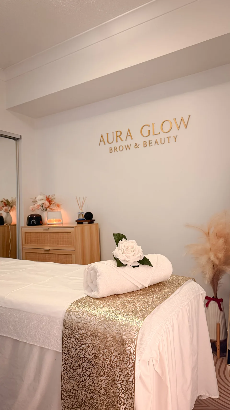 Aura Glow’s treatment bed with white linen, a rolled towel and gold runner beneath the Aura Glow Brow & Beauty wall sign